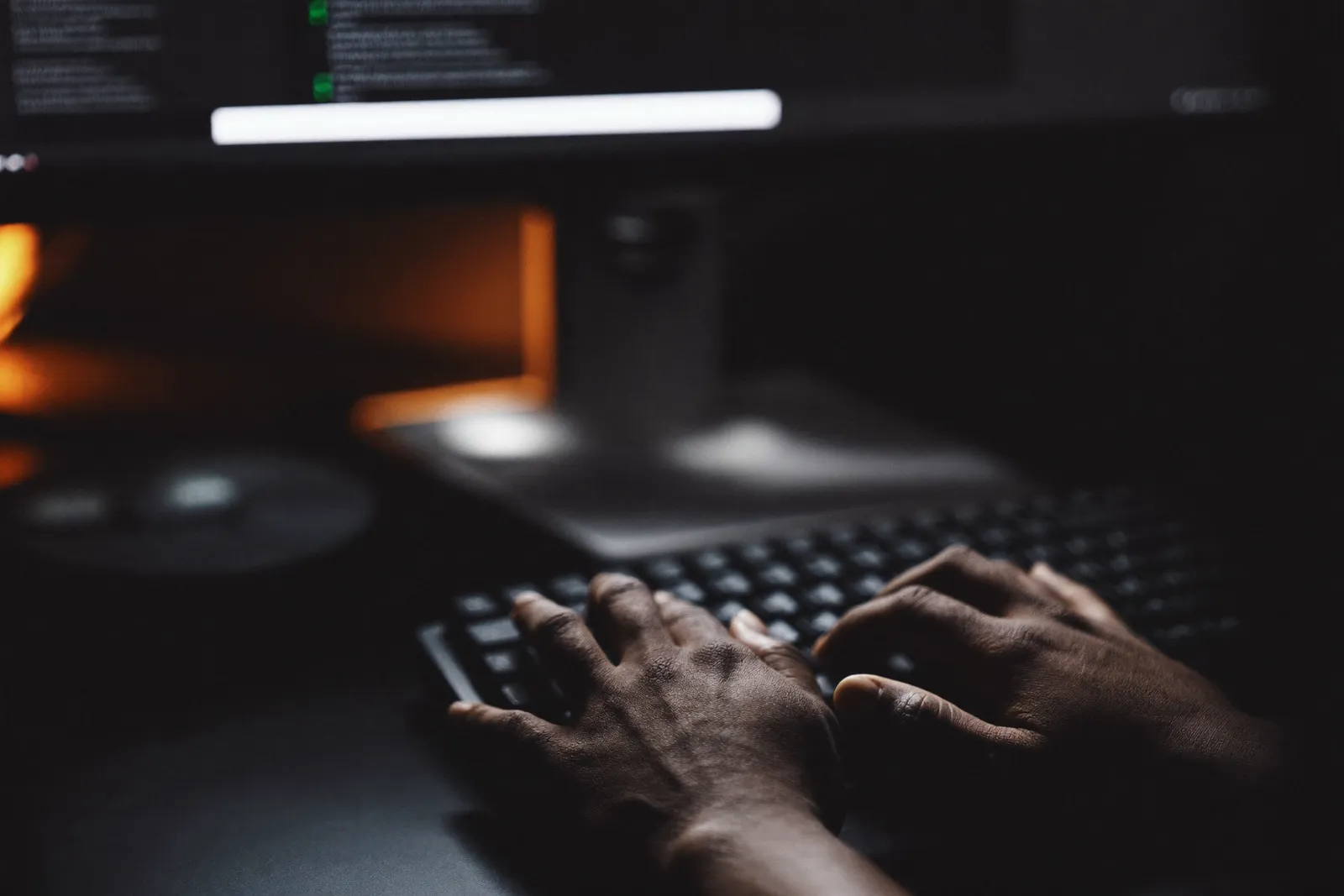 A developer's hands typing on a backlit keyboard, code on the screen behind.